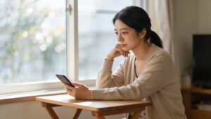 窓辺のデスクで朝の光のなか、スマホを静かに見つめる30代女性。落ち着いた表情で配偶者の行動を整理する場面。