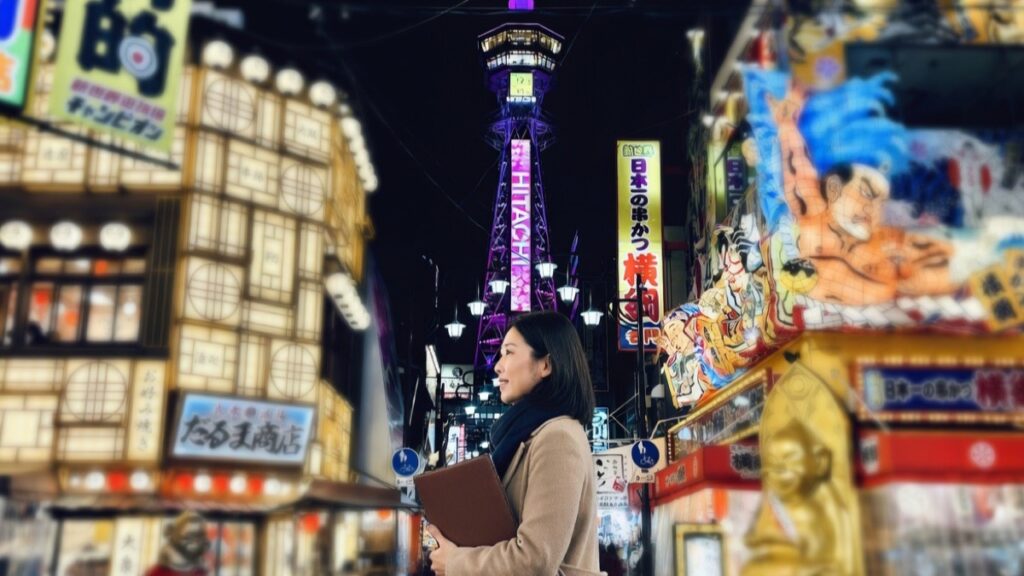 夜の新世界の通天閣を背景にベージュコートで革のドキュメントフォルダを抱える女性。大阪での探偵相談・調査を象徴するイメージ。