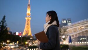 夕暮れの名古屋テレビ塔を背景にチャコールコートで革フォルダを抱える女性。愛知・名古屋での探偵相談・調査を象徴するイメージ。