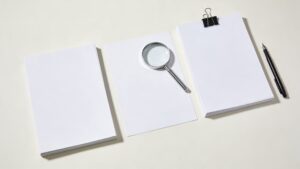 複数の白い書類束とシルバー虫眼鏡と万年筆のミニマル現代写真。戸籍調査と相続人探索を象徴。