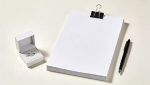 プラチナリングが入った白い指輪ケースと書類束と万年筆のミニマル現代写真。婚前の身辺調査と契約検討を象徴。