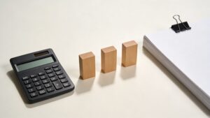 計算機・横並びの3本の木ブロック・書類束のミニマル現代写真。3つの料金プラン（時間制・成功報酬・パック）を比較する場面を象徴。